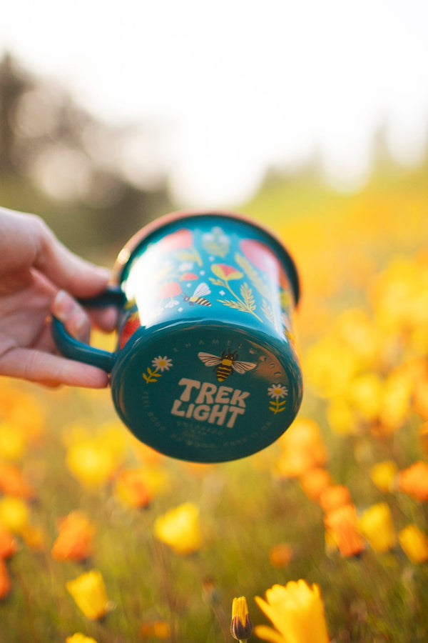 Colorful Trek Light Garden Buzz Enamel Mug 16oz in a field of orange flowers, perfect for outdoor adventures.