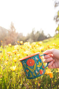 Garden Buzz Enamel Mug 16oz held in a field of yellow flowers, featuring vibrant floral design. Perfect for your next Trek Light adventure.