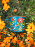 Trek Light 16oz Garden Buzz Enamel Mug amidst vibrant orange flowers, perfect for outdoor adventures.