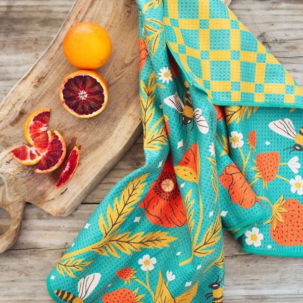 Garden Buzz kitchen and dish towel with vibrant garden design, eco-friendly and super absorbent, displayed with oranges on a wooden board.