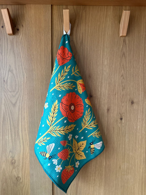 Eco-friendly Garden Buzz kitchen dish towel with vibrant floral and bee design, hanging on a wooden hook. Super absorbent.