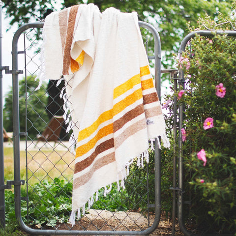 Handwoven Golden Age Blanket with brown and yellow stripes on a garden fence, made from 100% recycled materials, eco-friendly design.