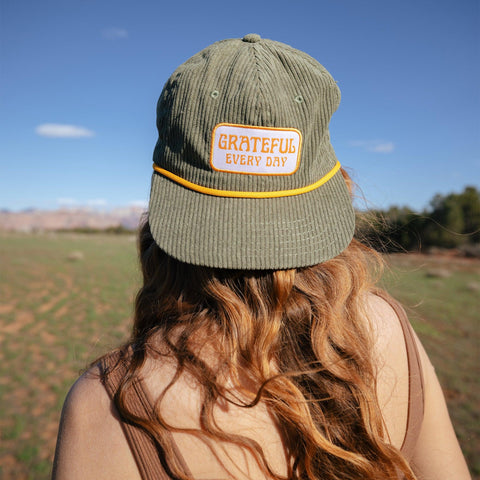 Woman wearing Grateful Every Day Trek Light corduroy snapback hat, enjoying nature with a green field background.