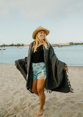 Woman wearing a Greyson Blanket at the beach, showcasing its stylish, handwoven design.