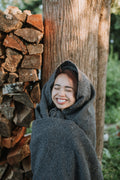 Happy woman wrapped in a Greyson Blanket near stacked firewood in a lush garden.