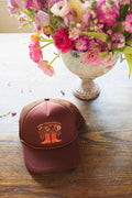 Grow Your Own Way Hat displayed on a wooden table with flowers, featuring a vibrant design and adjustable fit.