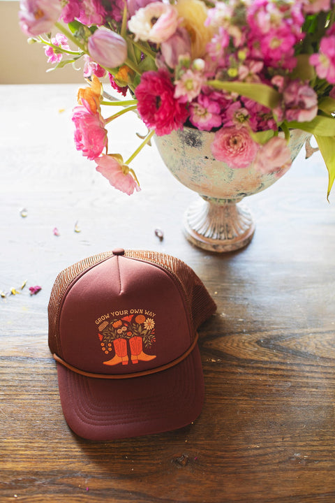 Grow Your Own Way Hat displayed on a wooden table with flowers, featuring a vibrant design and adjustable fit.