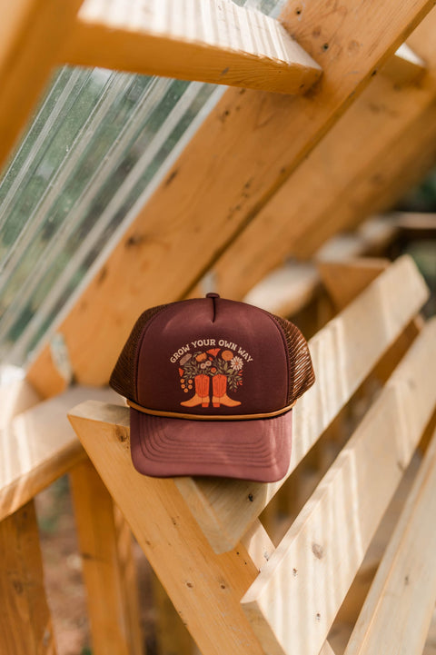 Grow Your Own Way Hat displayed on wooden shelf in greenhouse, featuring colorful design with flowers and boots.