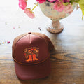 Grow Your Own Way Hat displayed on a wooden table with flowers, featuring a vibrant design and adjustable fit.