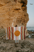 Woman holding a colorful striped blanket with a circular design, standing on a rocky beach. Trek Light inspired, handwoven beauty.