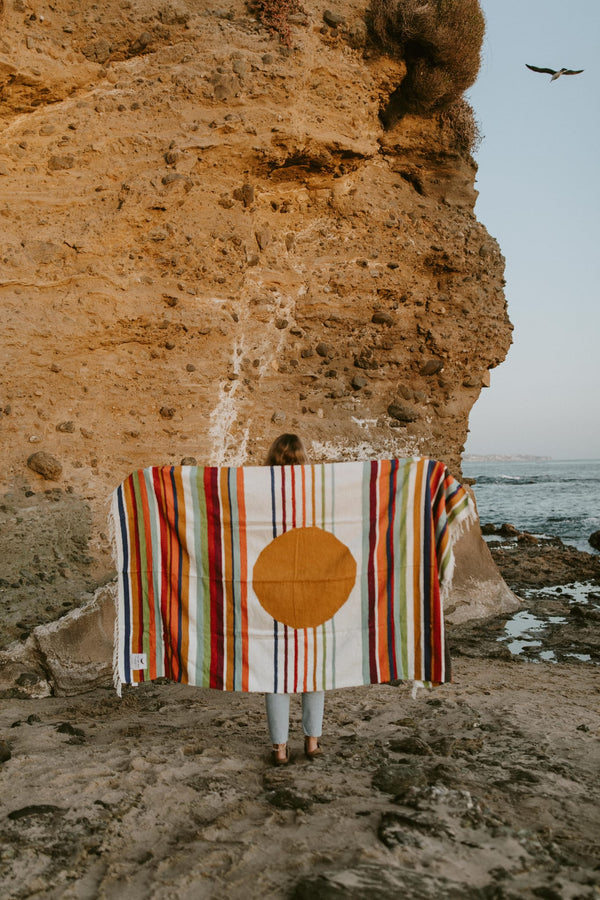 Woman holding a colorful striped blanket with a circular design, standing on a rocky beach. Trek Light inspired, handwoven beauty.