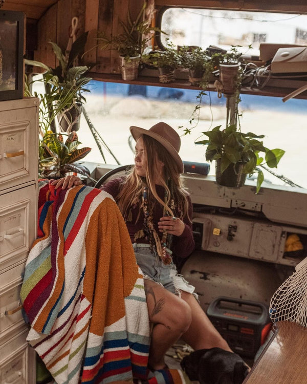 Cozy setting with a woman and a vibrant Halley's Comet blanket by Trek Light, surrounded by plants in a rustic space.