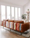 A brown leather sofa in a bright, modern living room, styled with a colorful striped handwoven Halley's Comet throw blanket draped over the seat.