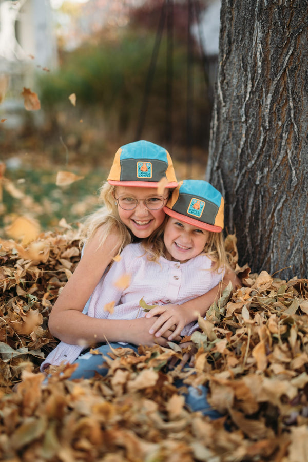 Two smiling children wearing Trek Light hats, sitting in a pile of autumn leaves near a tree. Perfect moment of outdoor joy.