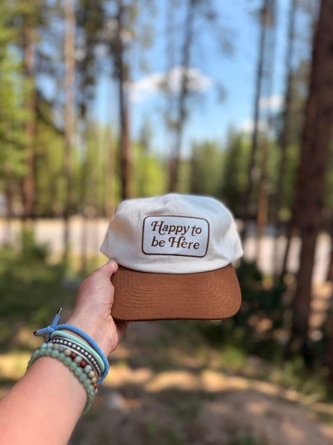 Happy To Be Here Hat held in hand, featuring a brown brim and comfortable design, perfect for all-day wear.