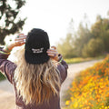 Back view of person wearing 'Happy To Be Here' hat amidst a sunny, flowered path, inspired by Trek Light vibes.