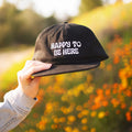 Trek Light black hat with 'Happy To Be Here' design, held against a vibrant outdoor background, showcasing joyful adventure vibes.