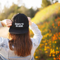 Person in a denim jacket wearing a 'Happy to Be Here' black cap, enjoying a scenic outdoor view surrounded by colorful flowers.