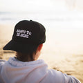 Person wearing a Happy To Be Here hat at the beach, expressing positivity and joy with Trek Light apparel.