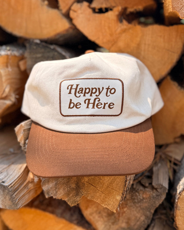 The Happy To Be Here Hat is a cream cap with a brown brim and a embroidered patch, resting on a stack of split firewood.