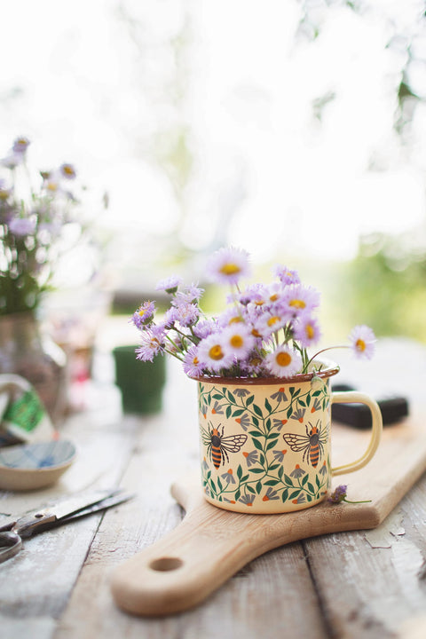 Honey Bee Enamel Mug 16oz filled with flowers on a wooden surface, perfect for adding charm to your mornings.