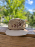 Howdy Hat displayed on a wooden surface with greenery in the background, featuring a flat-brim and adjustable fit.