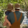 Father and child in matching 'Howdy' hats enjoy a serene nature walk by the riverside, embodying Trek Light adventures.