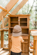 Child exploring wooden cabinet wearing Howdy Kids Hat, showcasing fun and adventure in outdoor play.