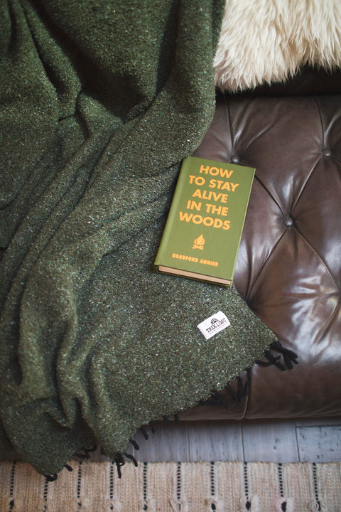Cozy Humboldt Blanket draped over a leather couch with 'How to Stay Alive in the Woods' book, handwoven and eco-friendly design.