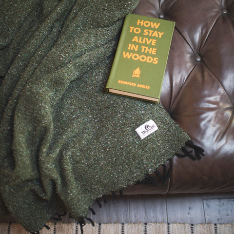 Cozy Humboldt Blanket draped over a leather couch with 'How to Stay Alive in the Woods' book, handwoven and eco-friendly design.