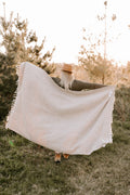 Woman holding a sand-colored Huntington Blanket in nature, perfect for beach days and cozy nights. Trek Light adventure vibes.