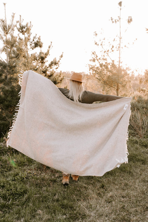 Woman holding a sand-colored Huntington Blanket in nature, perfect for beach days and cozy nights. Trek Light adventure vibes.