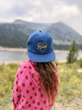A child viewed from behind wearing a blue denim baseball cap with 'Kiddo' embroidered in yellow inside a heart, paired with a pink strawberry-patterned hoodie, set against a blurred mountain and lake background.