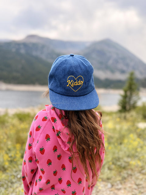 A child viewed from behind wearing a blue denim baseball cap with 'Kiddo' embroidered in yellow inside a heart, paired with a pink strawberry-patterned hoodie, set against a blurred mountain and lake background.
