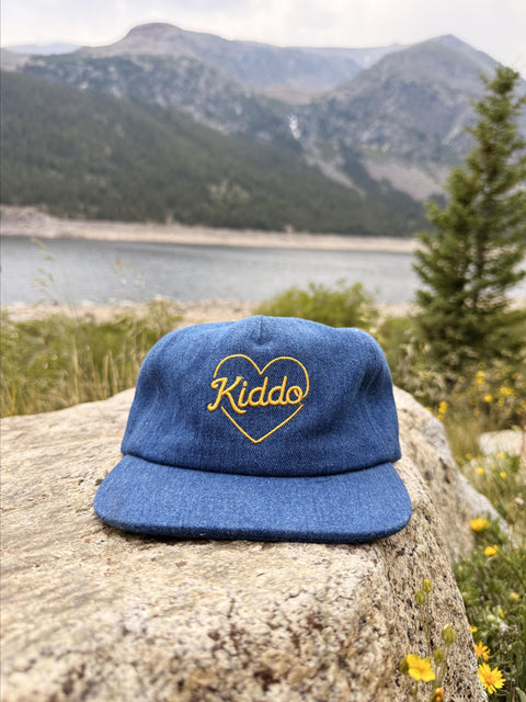 A blue denim-style flat-brim cap with a yellow embroidered heart and the word 'Kiddo' inside, resting on a large rock with a mountain lake landscape in the background.