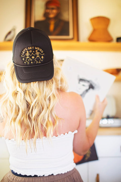 A woman wearing a black foam trucker hat with the phrase Let's Get Weird, showcasing a fun and unique style.