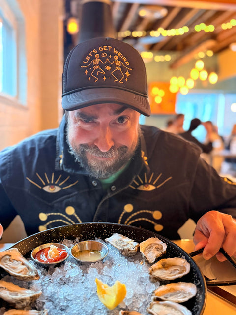 Man wearing a Let's Get Weird Hat, enjoying oysters at a vibrant restaurant, showcasing unique style.