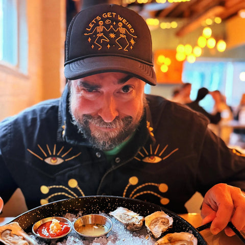 Man wearing a Let's Get Weird Hat, enjoying oysters at a vibrant restaurant, showcasing unique style.