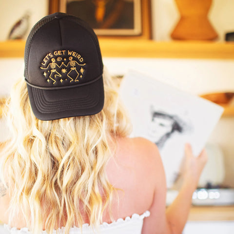 A woman wearing a black foam trucker hat with the phrase Let's Get Weird, showcasing a fun and unique style.