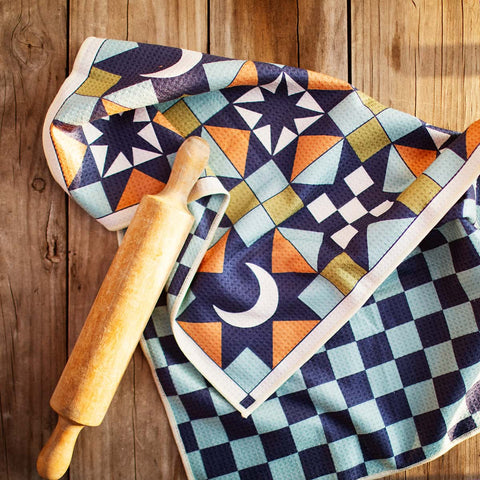 Colorful Trek Light kitchen dish towel with a stars and moon pattern, folded on a wooden surface with a rolling pin beside it.