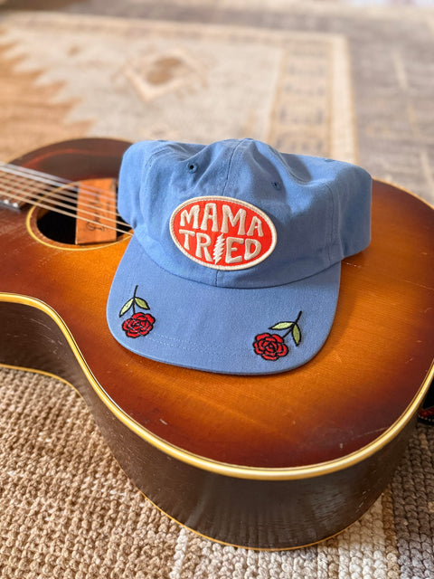 Mama Tried Hat displayed on a guitar, featuring vibrant red roses and an eye-catching logo.