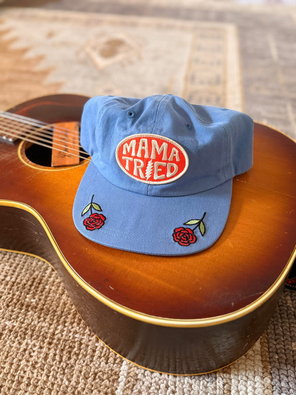 Mama Tried Hat displayed on a guitar, featuring vibrant red roses and an eye-catching logo.