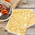 Yellow Meadow Kitchen & Tea Towel made from recycled materials, featuring a floral design on a wooden table next to a plate of citrus fruits.