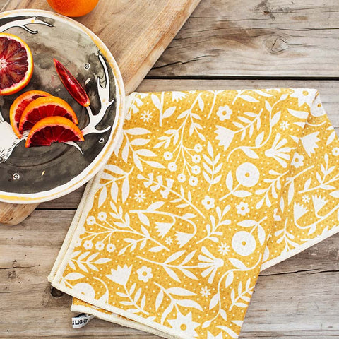 Yellow Meadow Kitchen & Tea Towel made from recycled materials, featuring a floral design on a wooden table next to a plate of citrus fruits.