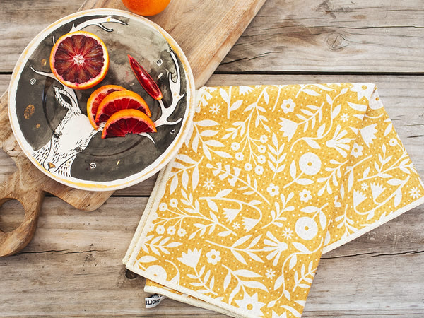 Yellow Meadow Kitchen & Tea Towel made from recycled materials, featuring a floral design on a wooden table next to a plate of citrus fruits.