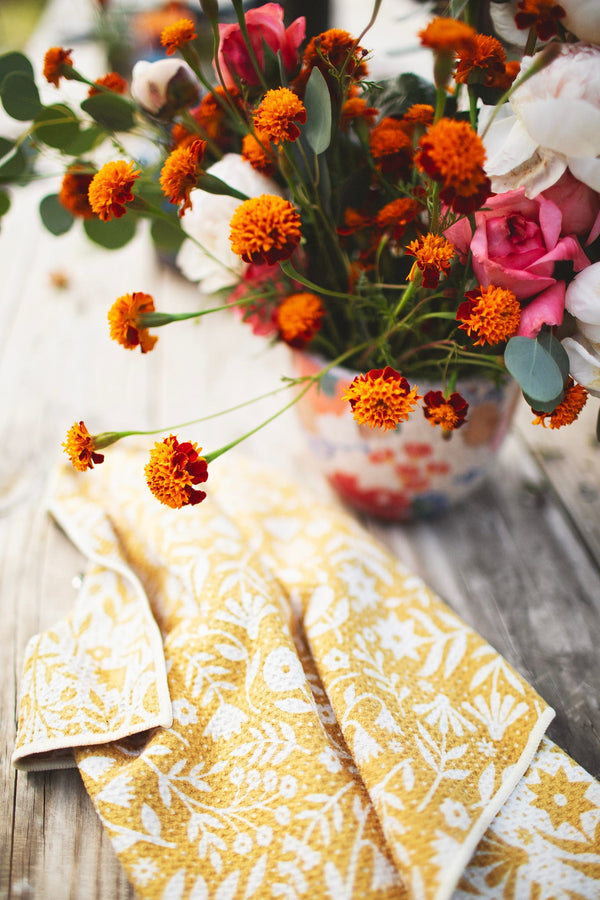 Meadow Kitchen & Tea Towel with vibrant floral design and hanging loop beside a flower pot.