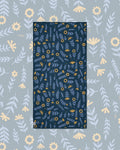Mermaids Wander & Travel Towel in navy with blue leaves and gold flowers, laid flat on a gray-blue floral background.