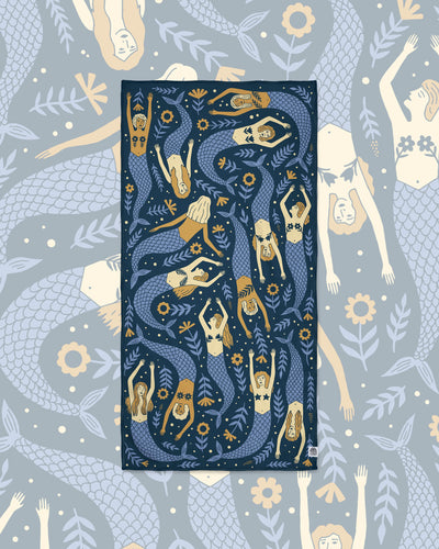 Mermaids Wander & Travel Towel in navy with blue-tailed mermaids and flowers, laid flat on a matching blue mermaid pattern.