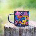 Midnight Mushroom Enamel Mug 16oz with colorful fungi design, resting on a wooden stump. Trek Light, perfect for outdoor adventures.