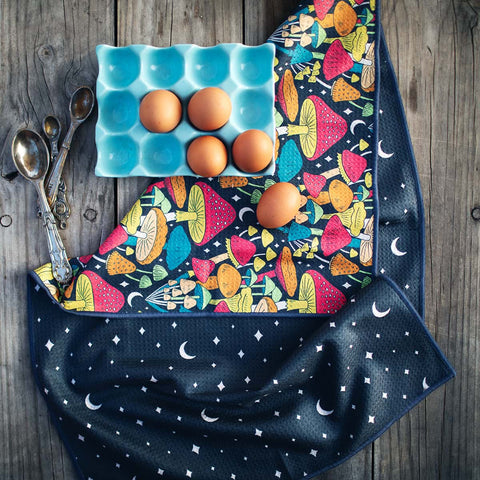 Midnight Mushroom Kitchen & Tea Towel with vibrant mushroom design, beside eggs in a blue tray and vintage spoons on a wooden table.